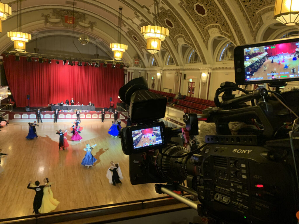 A Sony FX9 camera with a Canon CN7 lens on a tripod positioned to capture a ballroom dance scene in a grand, ornate hall. The TV production setup focuses on filming the dancers performing in vibrant costumes.