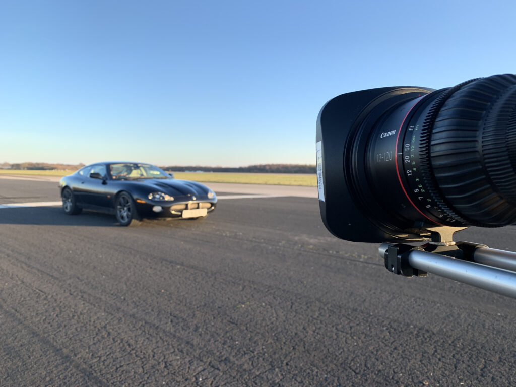 A black Jaguar sports car on the runway at Elvington Airfield in York, being filmed by a Sony camera with Canon CN7 lens during a video production. The outdoor setup aims to capture the car in motion with perfect lighting conditions under a clear blue sky for a promotional shoot for the charity Speed of Sight