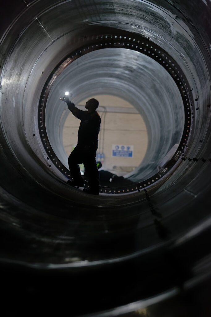 A worker inspects part of a large cylindrical piece of a wind turbine tower in a factory, with other sections of tower and machinery visible in the background.