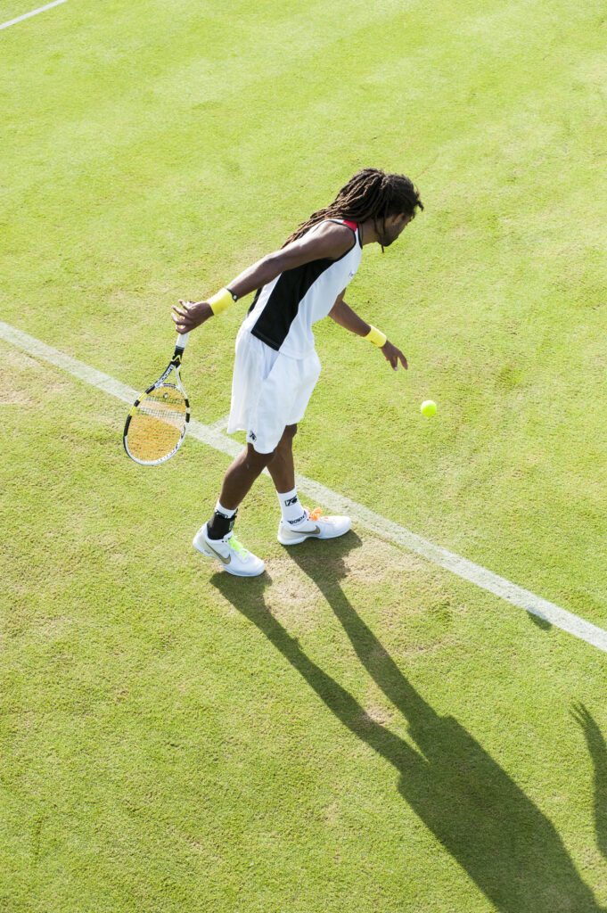A tennis player in a white outfit and yellow wristbands serves a ball on a grass court, viewed from overhead.