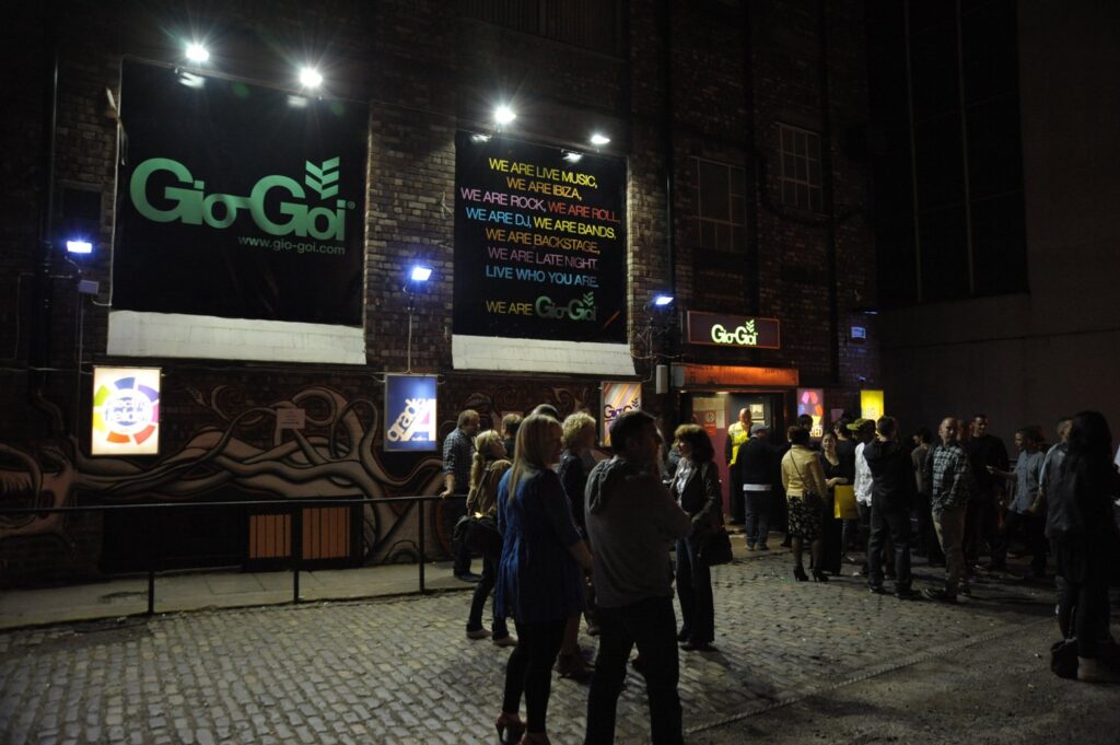 A group of people queuing outside a club with a GioGoi clothing brand banner. The building has art on the walls and illuminated neon signs.