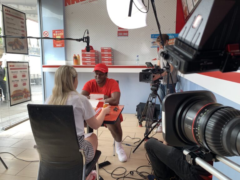 Behind-the-scenes of a video production in a fast food restaurant featuring a professional Sony camera / Canon CN7 lens combo capturing a chat between a person in a red top & his friend in white. Professional lighting equipment illuminates the scene, enhancing the quality of the production.